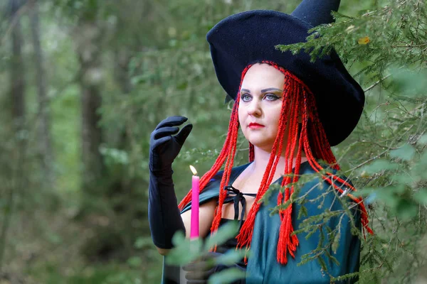 Das Gesicht der Zauberin mit roten Haaren in einem spitzen Hut und einem schwarzen Mantel im Wald in der Hand mit einer Kerze. — Stockfoto