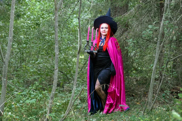 Eine Zauberin mit roten Haaren im Hut und einem violetten Mantel im Wald in der Hand mit einer brennenden Kerze. — Stockfoto