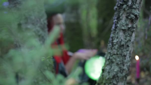 Una strega con i capelli rossi e un cappello nella foresta fa magia con una palla luminosa e candele accese — Video Stock