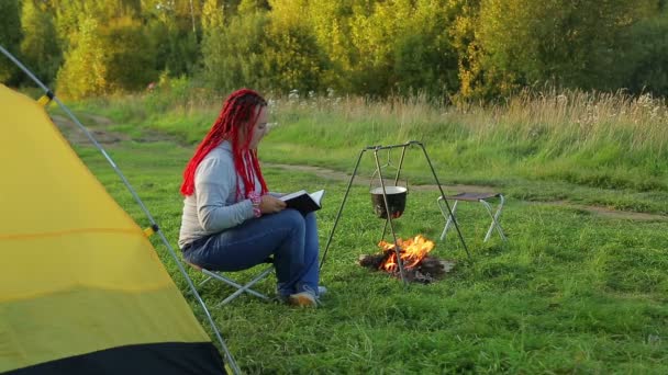 A woman near a campfire tent and watches a boiling pot. — Stock Video