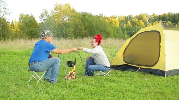 Un homme et une femme lors d'une soirée ensoleillée près d'une tente près d'un feu font dîner dans un pot . — Video