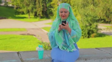 Parkta hafif bir eşarp genç bir Müslüman kadın kahve içiyor ve kulaklık ile haberci konuşuyor.