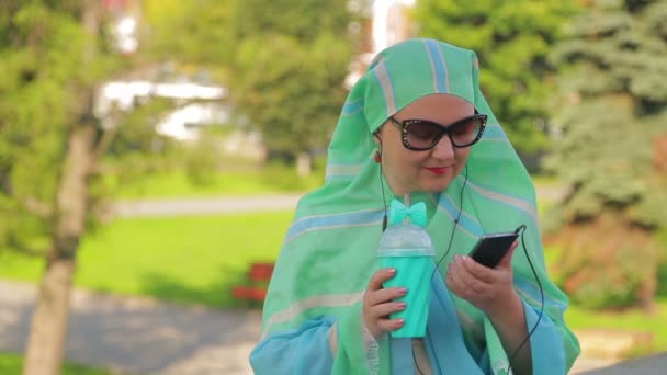 A young Muslim woman in a light scarf in the park is drinking coffee and talking in the messenger with headphones — Stock Video