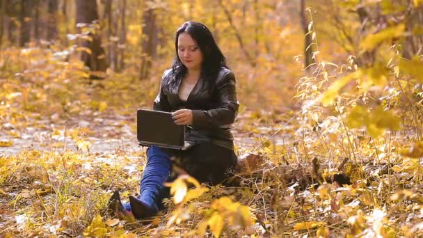Mladá brunetka v modrých botách sedí v parku za slunečného podzimního dne a pracuje na dálku s notebookem. — Stock video