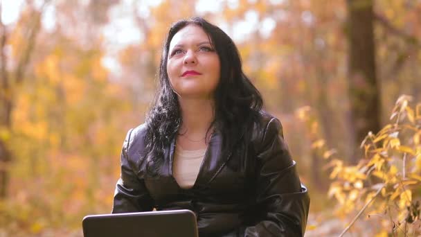 Mladá brunetka sedí v parku za slunečného podzimního dne, dívá se na obrazovku notebooku a usmívá se. — Stock video