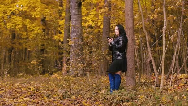 Μια νεαρή μελαχρινή γυναίκα με μπλε μπότες σε ένα πάρκο το φθινόπωρο στέκεται δίπλα σε ένα δέντρο και στέλνει μηνύματα στο τηλέφωνο. — Αρχείο Βίντεο