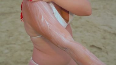 Beyaz mayo giymiş genç kızıl saçlı bir kadın deniz kıyısındaki cesede güneş kremi sürüyor..