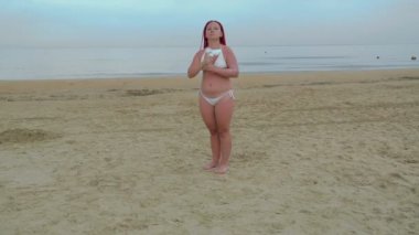 Beyaz mayo giymiş genç kızıl saçlı bir kadın deniz kıyısındaki cesede güneş kremi sürüyor..