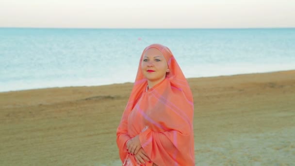 A pensive young Muslim woman in an orange shawl stands on the sand against the background of the sea. — Stock Video