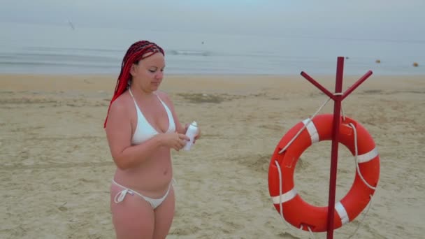 Una giovane donna in costume da bagno bianco applica la protezione solare sul corpo sulla riva del mare . — Video Stock