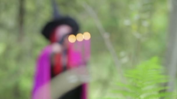 Fiatal boszorkány, vörös hajú, fekete kalapot és málnás köpenyt varázsol gyertyákkal az erdőben. — Stock videók