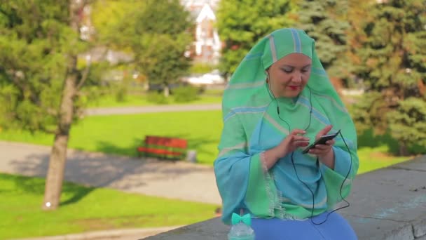 Una joven musulmana alegre en una bufanda ligera en el parque está bebiendo café y hablando en el mensajero con auriculares — Vídeos de Stock