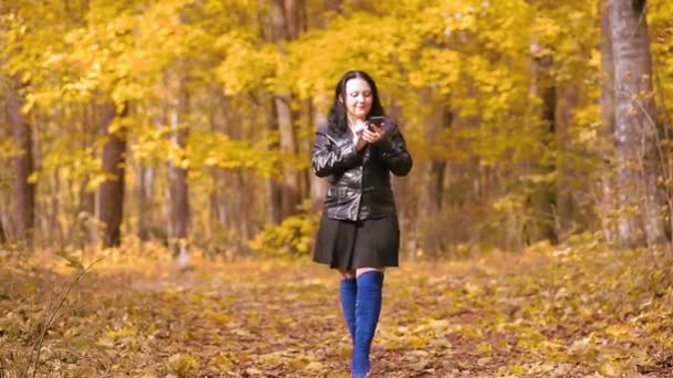 Μια νεαρή μελαχρινή με μπλε μπότες περπατάει στο πάρκο του φθινοπώρου με ένα τηλέφωνο στα χέρια της — Αρχείο Βίντεο