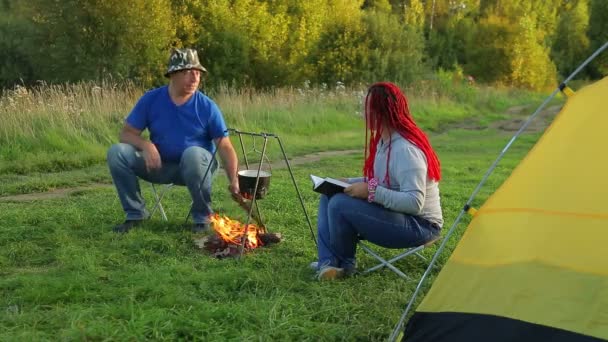 A man and a woman near a tent by the fire and watch a boiling pot. — Stock Video