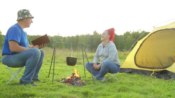 En man och en kvinna i en clearing nära ett tält nära en brand gör middag i en pott — Stockvideo