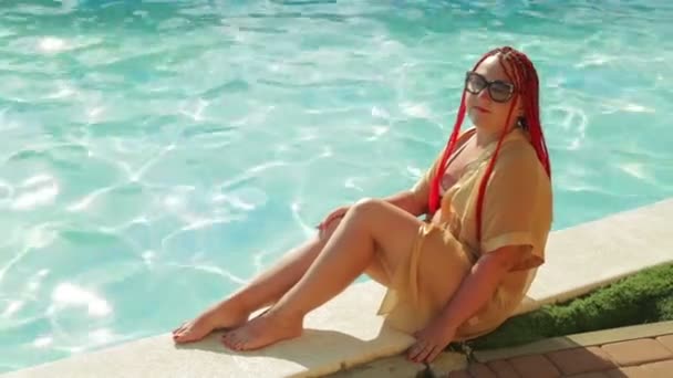 A young woman in a bathrobe and swimsuit is sitting on the edge of the pool — Stock Video