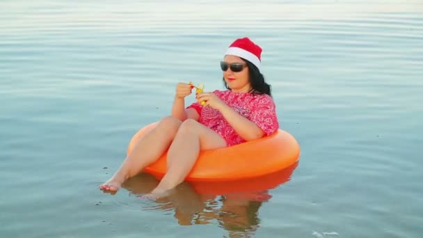 A brunette woman in a swimming circle and a Santa Claus hat eats a banana holds Christmas and New Year at sea — Stock Video