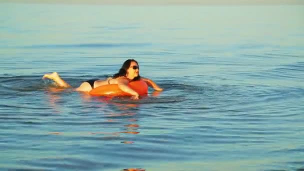Μια μελαχρινή γυναίκα με μαγιό κολυμπάει σε έναν κύκλο κολύμβησης στη θάλασσα κοντά στην ακτή. — Αρχείο Βίντεο