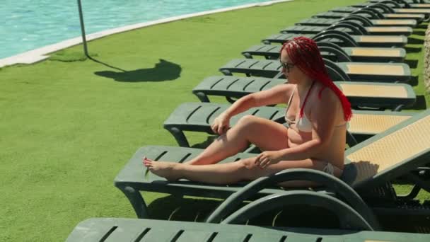 Eine moderne Juwelin im Urlaub am Pool in einer Sonnenliege beschmiert ihren Körper mit Sonnencreme — Stockvideo