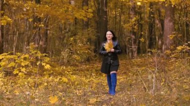 Mavi çizmeli esmer bir kadın elinde sarı yapraklarla sonbahar parkında yürüyor.