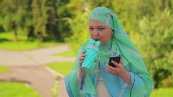 Una joven musulmana en una bufanda ligera en el parque está tomando café y hablando en el mensajero con auriculares . — Vídeos de Stock