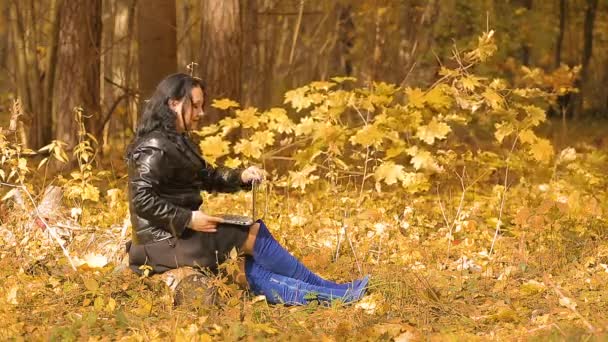 Eine brünette Frau in blauen Stiefeln sitzt an einem sonnigen Herbsttag in einem Park und arbeitet aus der Ferne mit einem Laptop — Stockvideo