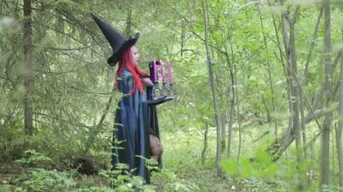 kızıl saçlı genç cadı ve siyah bir şapka ve siyah pelerin ormanda mumlar ile conjures.