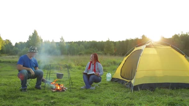 En man och en kvinna läser en bok nära ett tält nära en lägereld och tittar på en kokande gryta. — Stockvideo