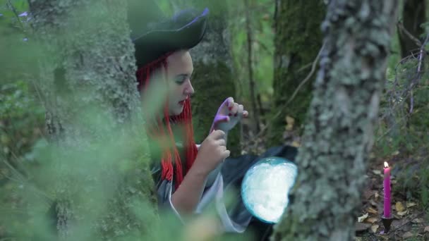 A sorceress with red hair and a hat in the forest does magic with a luminous ball and burning candles — Stock Video