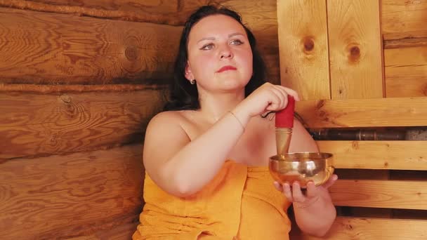 Une jeune femme brune dans un bain dans une serviette médite avec un bol chantant . — Video