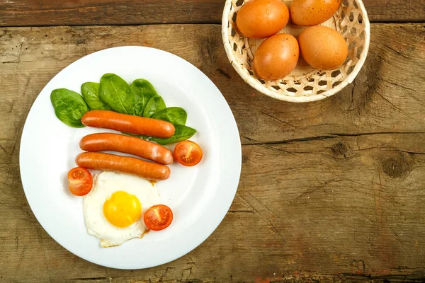 White plate with fried egg with sausages and tomatoes on a wooden table. — Stock Photo, Image