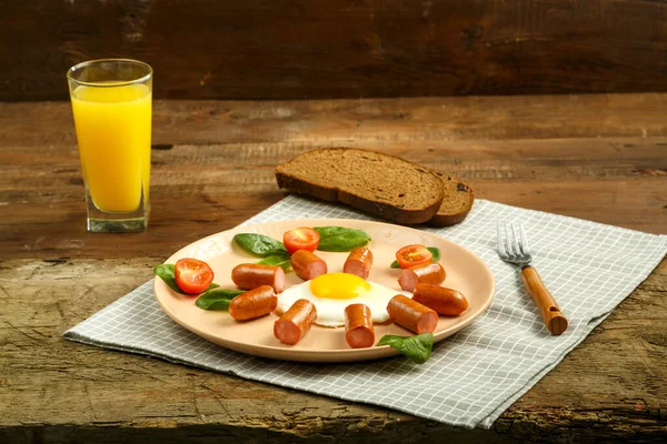 Prato de café da manhã com ovos fritos com linguiças e tomates em uma mesa de madeira e suco . — Fotografia de Stock