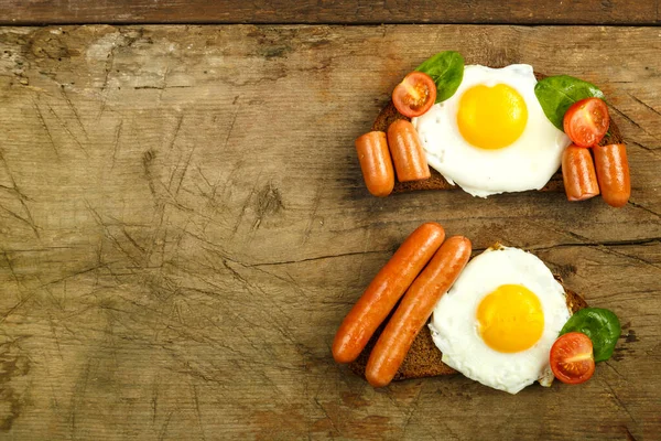 Duas sanduíches Ovos fritos com linguiças e tomates com ervas em um pedaço de pão em uma mesa de madeira . — Fotografia de Stock