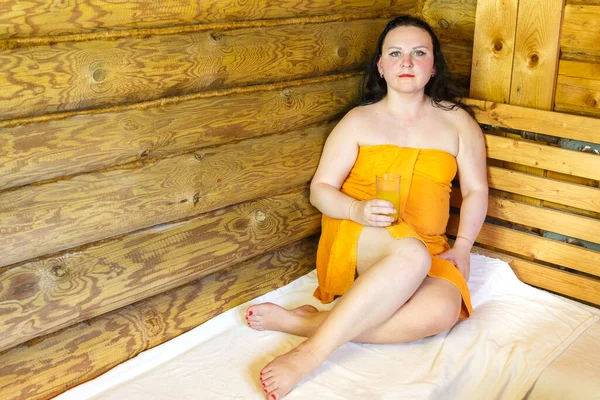 Een brunette vrouw in een bad gewikkeld in een handdoek zit met een glas sap. — Stockfoto