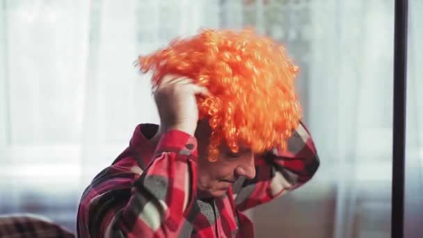 En man framför en spegel sätter på sig en clownperuk med röda lockar — Stockvideo