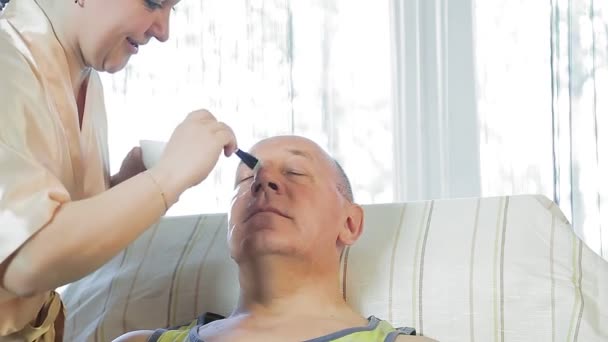 Una mujer en casa aplica una máscara a la piel de un hombre . — Vídeos de Stock