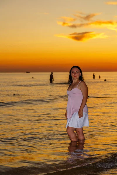 En ung kvinna i vit klänning står på stranden vid solnedgången.. — Stockfoto