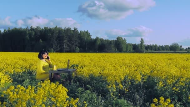 En kvinna i gula kläder i mitten av ett fält med gula blommor arbetar på distans vid en dator och dricker juice och talar i telefon — Stockvideo