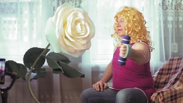 En transvestit i en paryk med make-up derhjemme ryster håndvægt biceps. – Stock-video