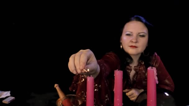 En spåkvinna i en magisk salong tänder ett ljus med en tändsticka på en svart bakgrund. — Stockvideo
