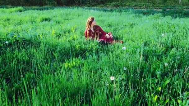 En rödhårig zigenare i röd klänning kopplar av på gräset på ett fält. — Stockvideo