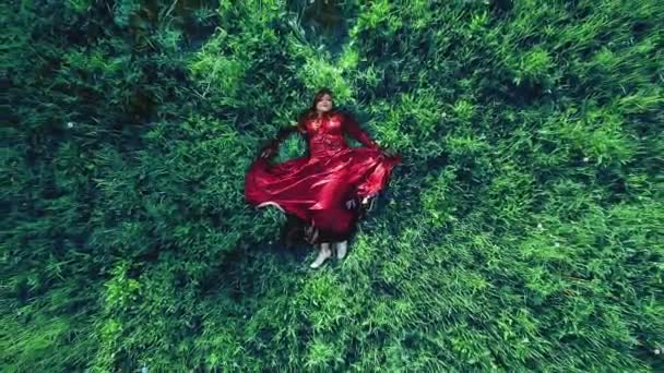 Μια νεαρή τσιγγάνα με κόκκινο φόρεμα ξαπλώνει στο γρασίδι σε ένα χωράφι.. — Αρχείο Βίντεο