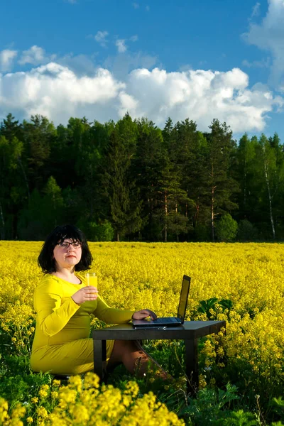 Молода жінка фрілансер в полі серед жовтих квітів за столом п'є сік . — стокове фото