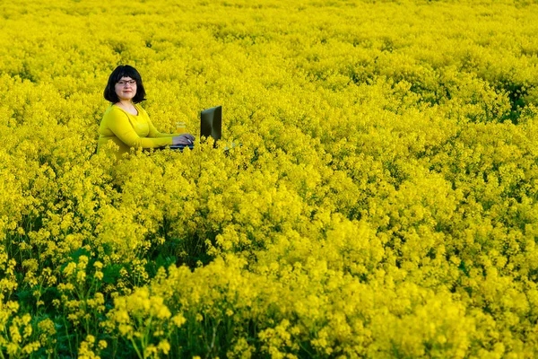 Ung kvinna frilansare i ett fält bland gula blommor vid ett bord med och en dator. — Stockfoto