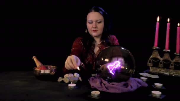 Ένας μάντης σε ένα μαγικό κομμωτήριο ανάβει κεριά σε μαύρο φόντο πίσω από μαγική φωτιά. — Αρχείο Βίντεο