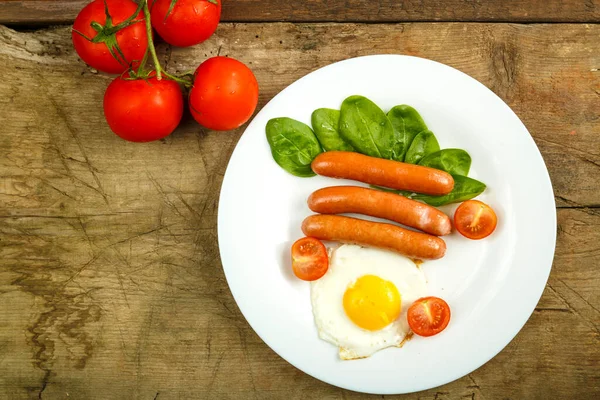 White plate with fried egg with sausages and tomatoes on a wooden table. — Stock Photo, Image