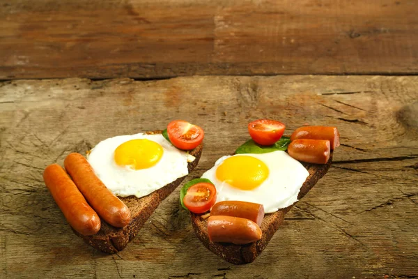 Duas sanduíches Ovos fritos com linguiças e tomates com ervas em um pedaço de pão em uma mesa de madeira . — Fotografia de Stock