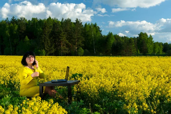 Młoda kobieta freelancer na polu wśród żółtych kwiatów przy stole pije sok. — Zdjęcie stockowe