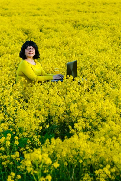En ung kvinna arbetar på ett fält bland gula blommor vid ett bord med en bärbar dator. — Stockfoto