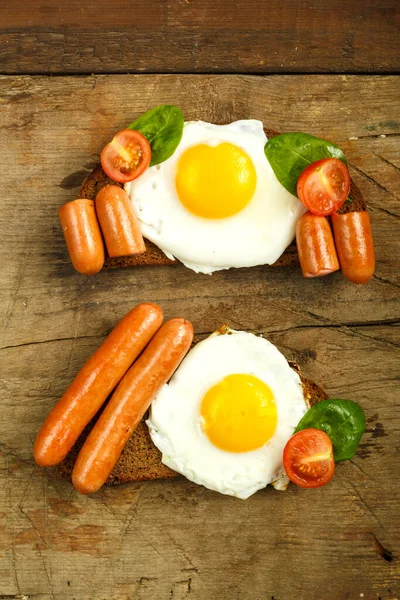 Duas sanduíches Ovos fritos com linguiças e tomates com ervas em um pedaço de pão em uma mesa de madeira . — Fotografia de Stock